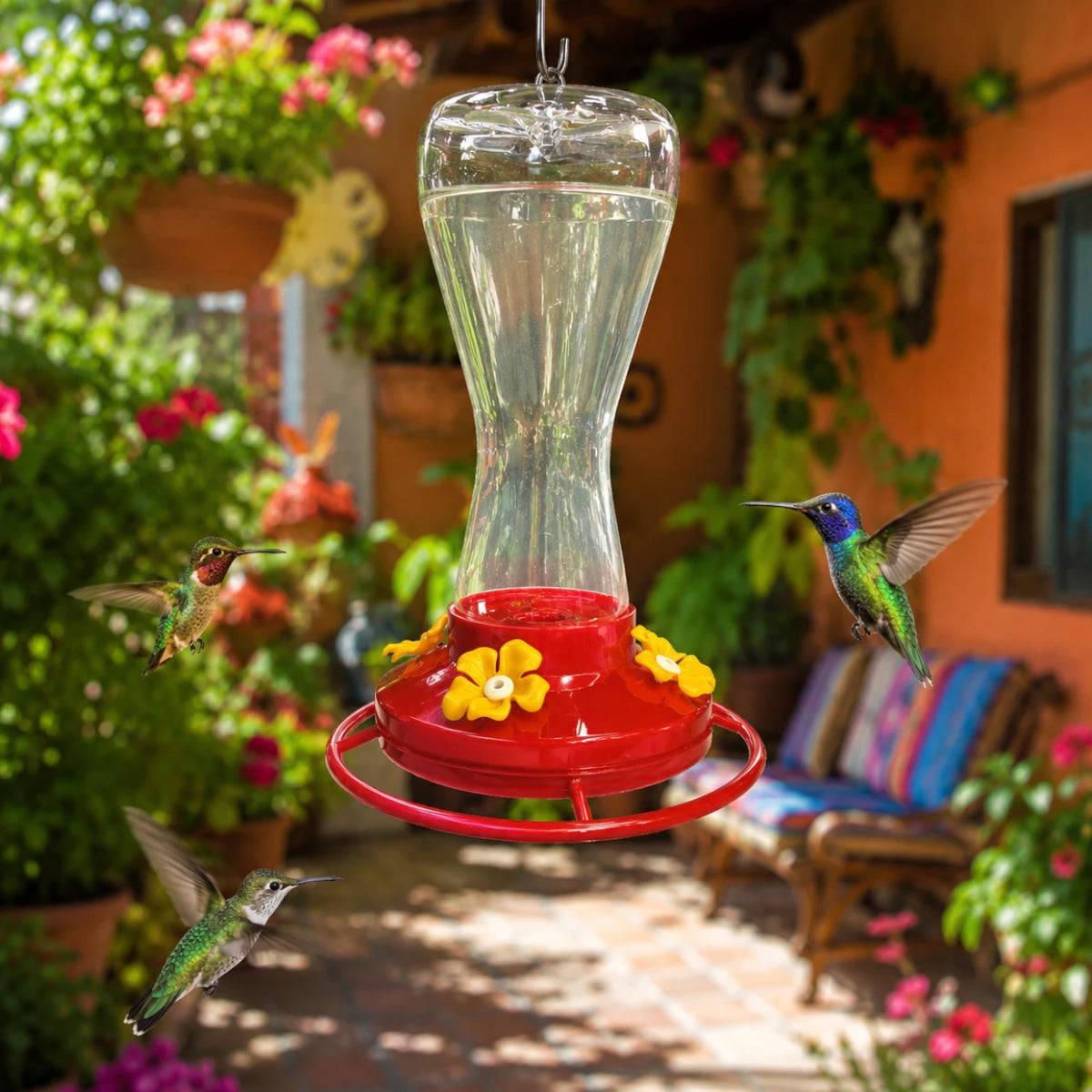 Tres colibríes revoloteando alrededor del bebedero Redkite de 470 ml colgado en una terraza iluminada con plantas y arbustos de fondo, en Kamalitas.