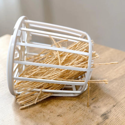 Dispensador de yute tipo canasta plástica en color blanco, lleno de fibras naturales y listo para su uso en jaulas de aves, en Kamalitas.
